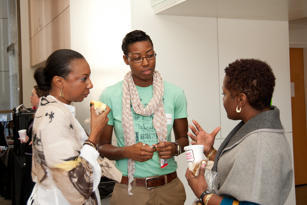 Three women are standing and talking in a building. The walls behind or white. In the distance you can see some light wood wall covering that is contemporary. The woman in the center is wearing a mint green T-shirt and a white scarf with a brown belt and khakis. The woman on the left is wearing a white blouse with a black abstract design on it. The woman on the right is wearing a gray top which folds over at the collar to reveal a dark gray. She is revealing the left side of her face which shows one weighty silver hoop earring. She is holding a cup in her left hand. All three are engrossed in discussion. All three women are dark skinned.