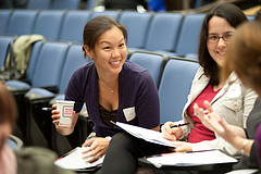 Three Women sitting in blue chairs in what appears to be an auditorium are leaning over and talking. One woman is holding a drink wearing a black sweater and smiling. She is Asian. She is in the center of the photograph. A Caucasian woman with dark hair and glasses, wearing a red T-shirt with a white jacket. It's to the right of the photo and also smiling. They are both looking at a third person, we see only her light brown hair from the back. They all have folders in their laps and pens in their hands.