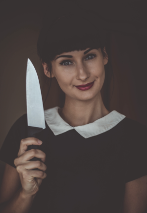 A woman with black hair that is pulled back and bangs is smirking and holding a large knife upwards, about 2 inches to the side of her head as if on display. She is wearing a black top with a white collar, which looks pretty puritanical. Her hair is back and she has thick Black bangs. She is Caucasian with olive skin, and has on red lipstick, and very well defined eyebrows. She looks like somebody's friendly mom or neighbor, other than the fact that she is holding this big knife.